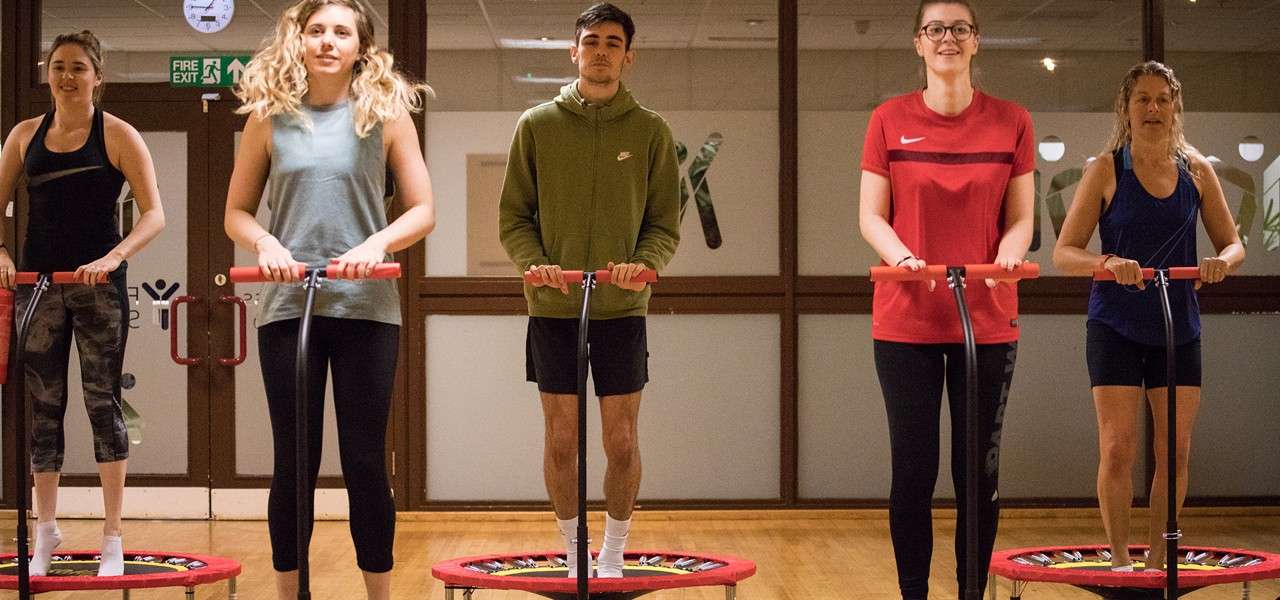 Five adults exercise, bouncing on handlebar mini-trampolines, facing forward. In a gym studio with wooden floor and glass doors behind. Text seen: FIRE EXIT.