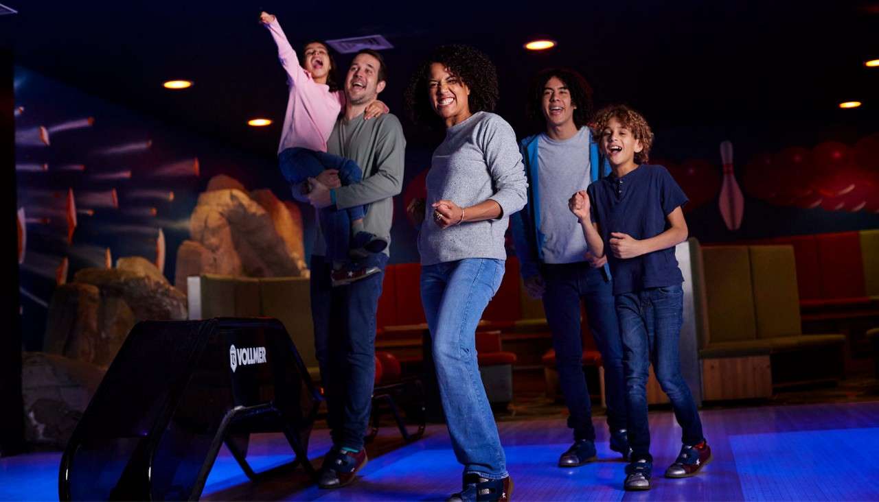 Family of five celebrates, smiling and cheering after a bowling roll; one adult holds a child, others in bowling shoes watch; neon-lit bowling alley with benches. Text: VOLLMER.