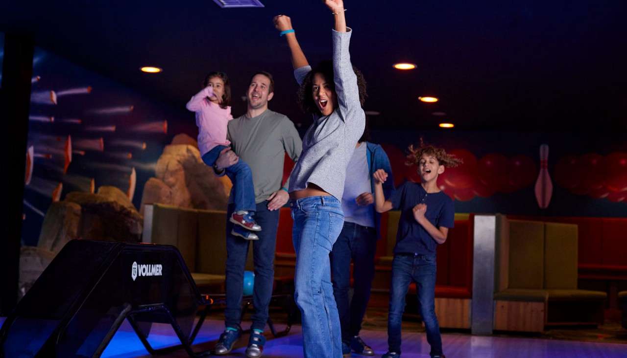 Family group celebrates, raising arms and cheering; a man holds a child. They stand near a bowling lane and ball return under dim, colorful lights. Text: 13 VOLLMER.