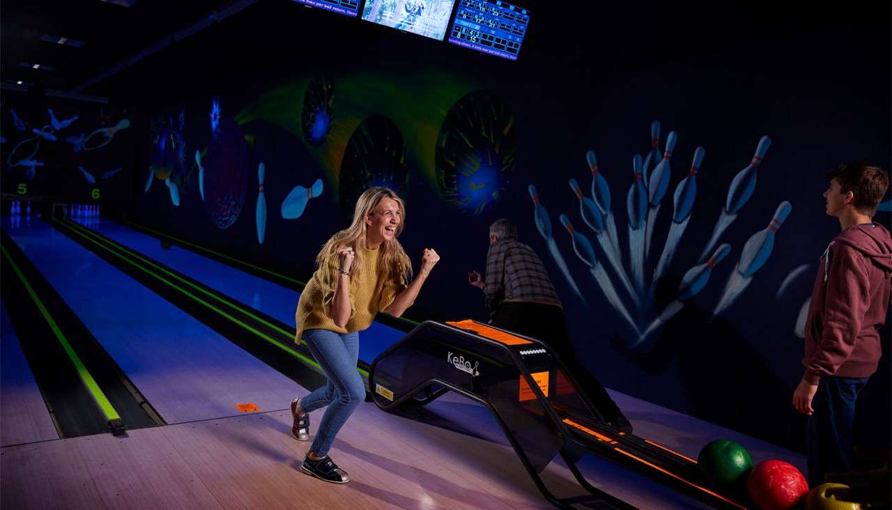 Woman celebrates a bowling roll, pumping her fists; companions watch. Neon-lit lanes lead to pins with wall art of flying pins. Visible text: lane numbers 5, 6.
