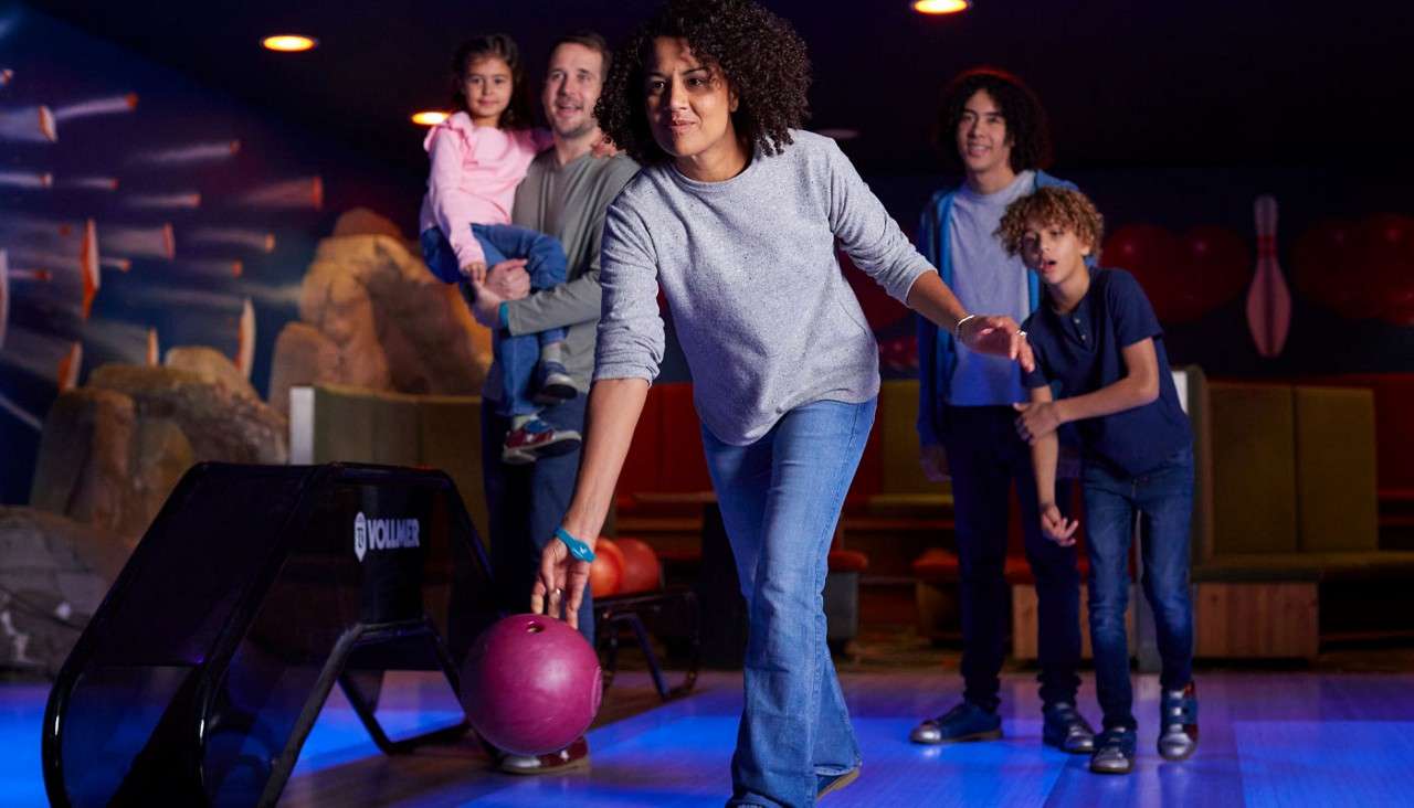 Woman bowls, swinging a pink ball toward the lane, while family members watch behind—man holding a small child and two boys. Indoor bowling alley with mural and seating. Text: “VOLLMER”.