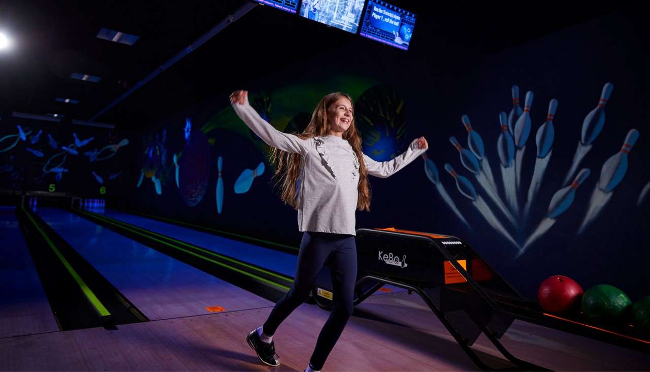 Girl celebrates with arms out, stepping off a bowling ramp. Neon-lit bowling alley surrounds her with mural pins, overhead screens, balls, lanes, and lane numbers "5." Visible text: "KeBoL", "5".