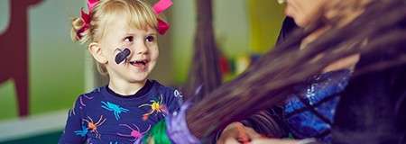 Child with face paint and pink hair bows smiles at a costumed adult holding a broom, in a colorful indoor playroom with soft lighting and playful decorations.