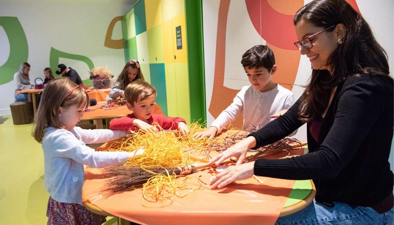 Children and an adult craft with straw and twigs, arranging materials on an orange table, in a bright, colorful workshop room where other groups work at nearby tables.