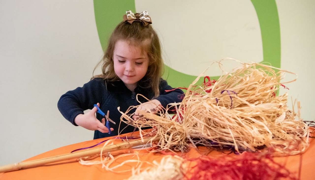 Young child cuts raffia strands with blue scissors at an orange-covered table. A bundle of straw-like fibers and a bamboo stick lie before her, indoors.