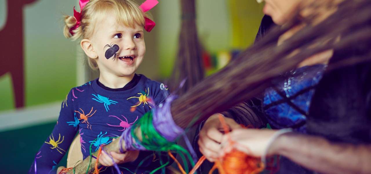 Smiling child with spider face paint and ribboned pigtails watches an adult wrap colorful yarn around a broomstick during a playful indoor craft activity.