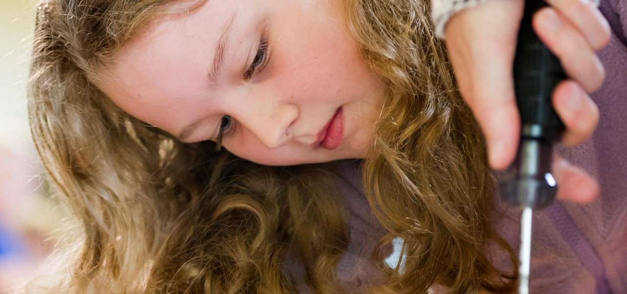 Child concentrates while using a screwdriver on a project; warm indoor lighting and a shallow depth of field blur the background, suggesting a workshop or classroom setting.