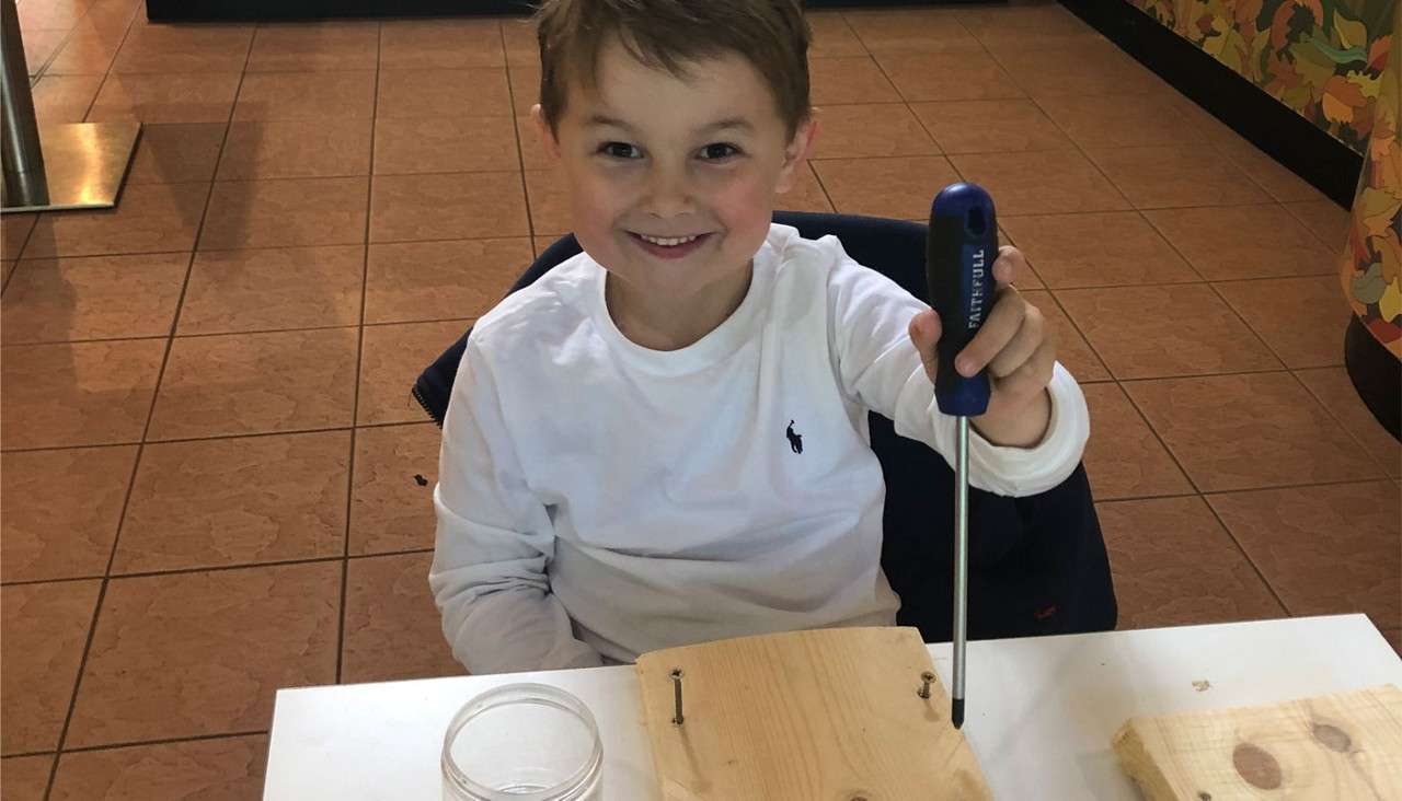 Child holds a blue-handled screwdriver and tightens screws into a wooden board, seated at a white table in a tiled room; a small jar sits nearby. Text on tool: “FAITHFULL.”