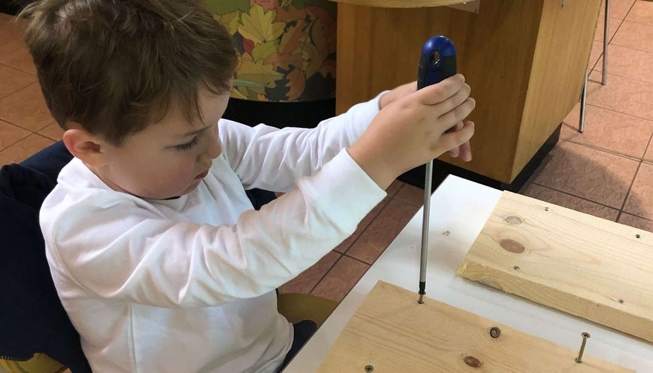 Child uses a long screwdriver to drive a screw into a wooden board at a table, with extra boards and screws nearby in an indoor workshop or classroom.