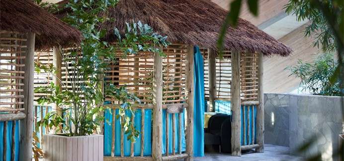 Thatched wooden cabanas stand side-by-side, their slatted walls and turquoise curtains framing cushioned seating, while potted tropical plants surround them in a bright, modern indoor lounge with stone surfaces.