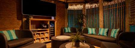 Curved wicker sofas encircle low round tables, inviting relaxation; in a tiki-style lounge with bamboo walls, teal-striped cushions, a large TV, shelf of rolled towels, potted plant, slatted windows.