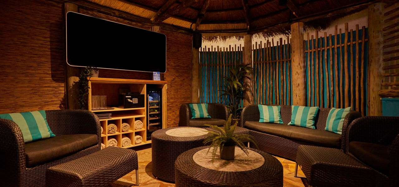 Wicker sofas encircle round tables, creating seating. In a cozy thatched-roof cabana, teal-striped pillows, a blank TV, shelving with rolled towels, safe, mini-fridge, and plants sit against wooden slat walls.