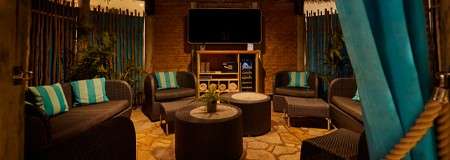 Wicker lounge seating encircles two round tables, hosting a small plant, in a covered cabana; a wall-mounted TV and shelves stand ahead, amid bamboo walls, plants, and turquoise curtains.