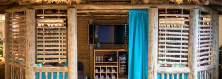 Cabana shows a TV and shelves with glasses and rolled towels, inside a rustic thatched hut, flanked by slatted wooden windows and a turquoise curtain, suggesting a relaxed poolside setting.