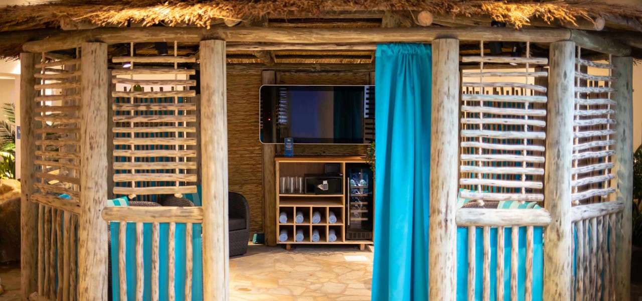 Thatched wooden cabana reveals an interior with wall-mounted TV, shelving holding glasses, rolled towels, and mini-fridge; wicker seating with striped cushions sits on a stone floor.