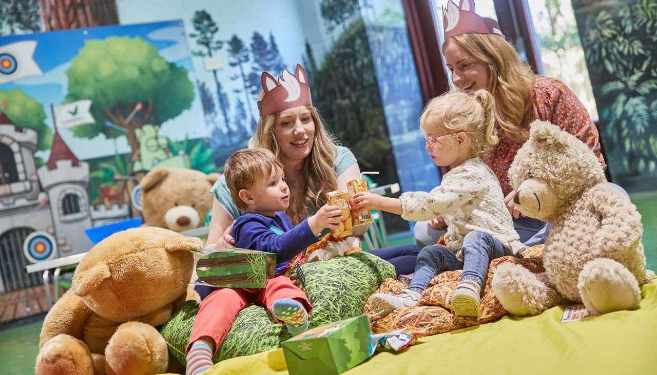Two children toast juice boxes while sitting on cushions, flanked by large teddy bears. Two adults wearing animal-ear paper crowns watch, in a playroom decorated with forest and castle murals.