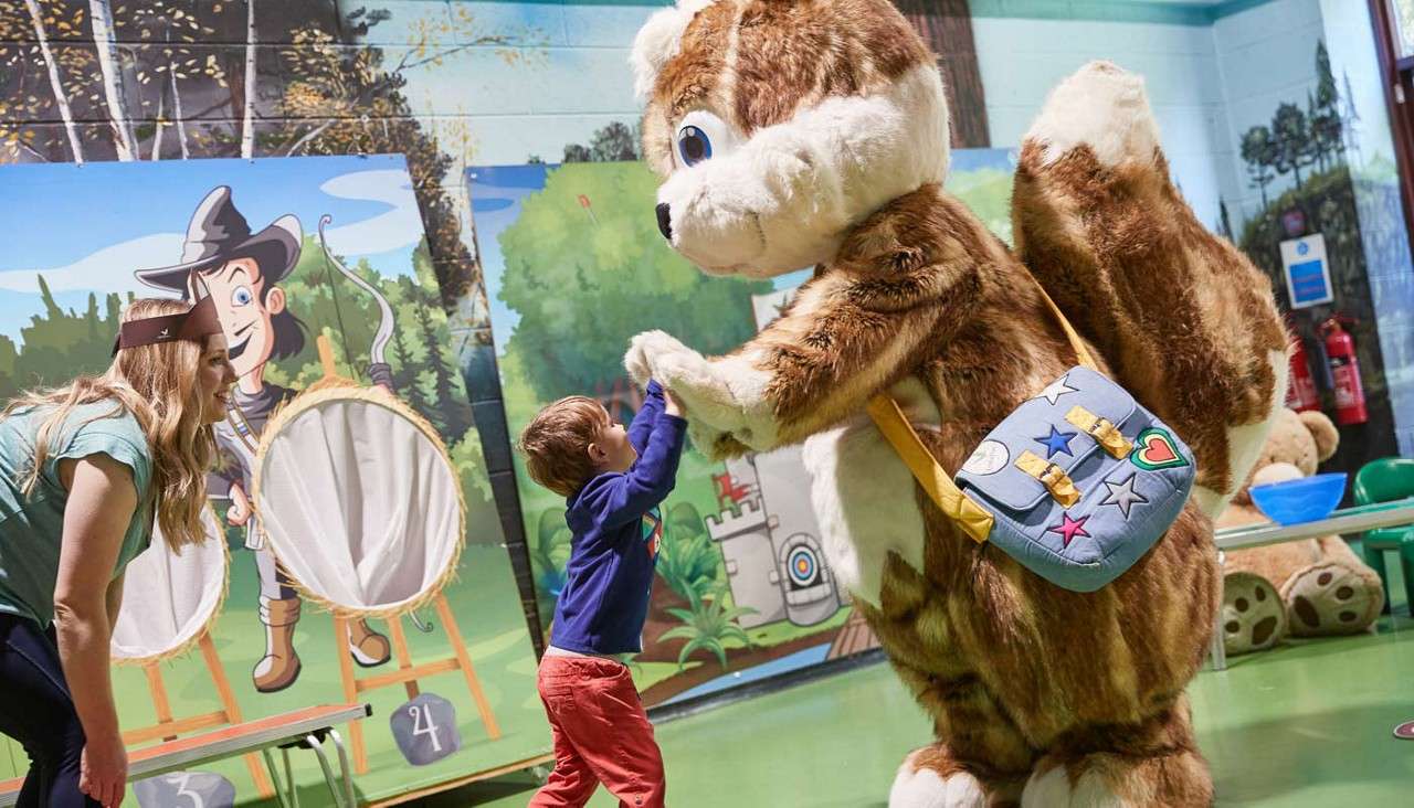 Large squirrel mascot high-fives a young boy, while a woman watches in an indoor play area decorated with forest-themed murals and archery targets; green floor, toys and chairs around.