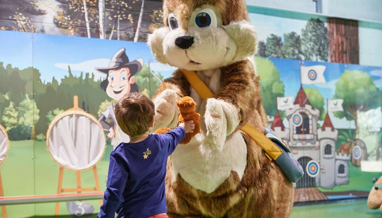 A person in a chipmunk mascot costume receives a teddy bear from a child, in a play area with cartoon forest and castle murals, archery targets, and a floor marking "3".