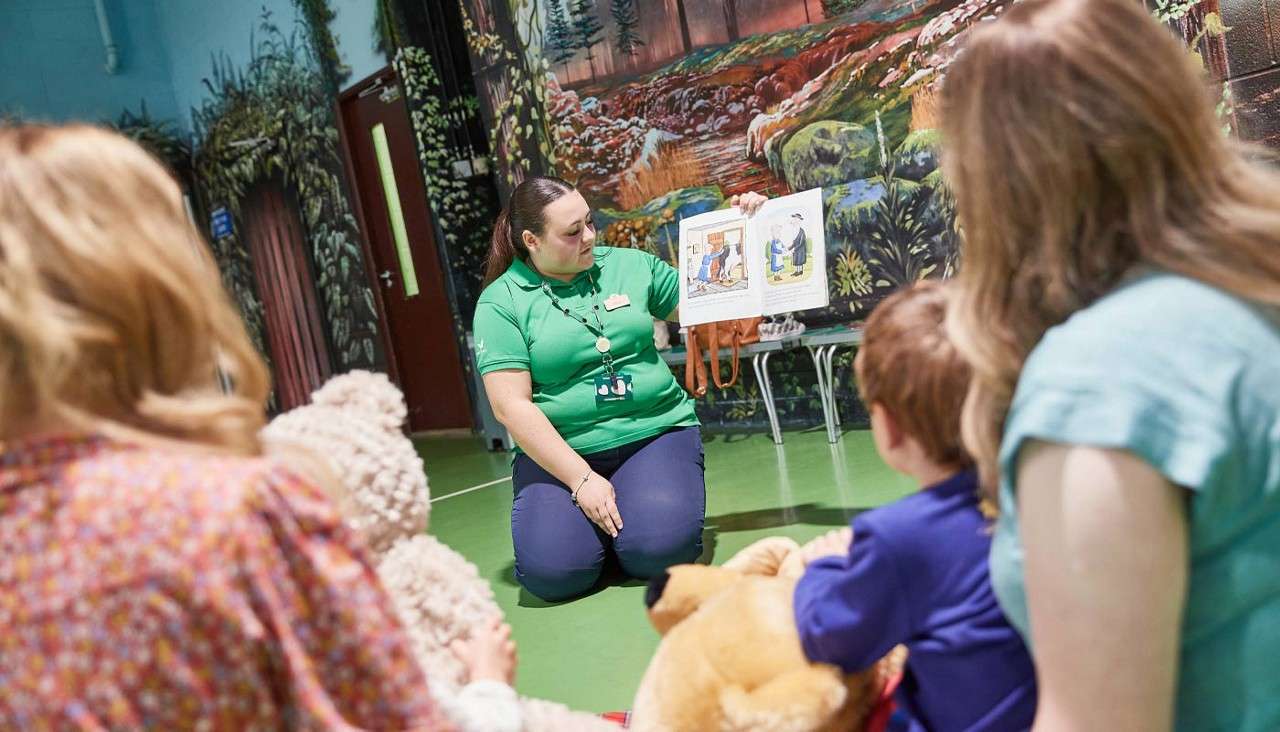 Teacher holds an open picture book, reading to children seated with teddy bears, in a gym-like room with forest mural walls; the small audience watches attentively during group storytime.