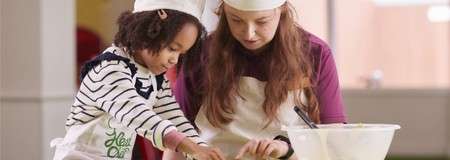 An adult and a child wearing chef hats and aprons knead dough together at a table; a mixing bowl and utensils nearby in a bright indoor cooking class. No legible text.
