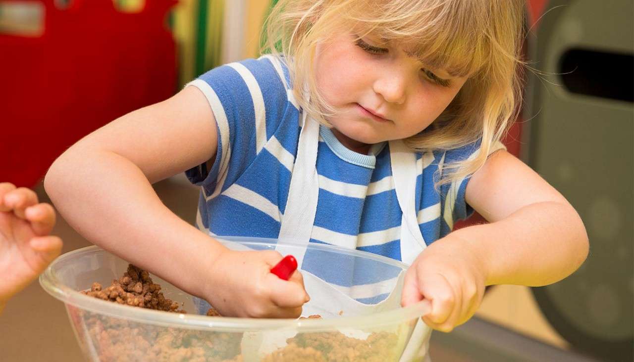 Child stirs mixture in a large plastic bowl using a red-handled utensil, wearing a striped shirt and apron, in a bright indoor setting, another child's hand visible nearby.