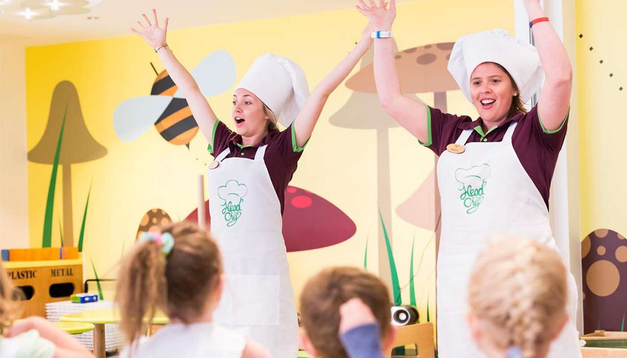 Two instructors in chef hats raise their arms, leading children in an activity, in a colorful classroom with mushroom and bee murals; aprons read "Head Chef." Recycling bin labeled "PLASTIC METAL".
