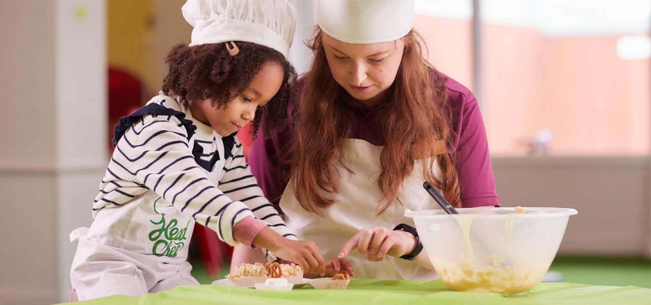 Child in chef hat decorates cupcakes with an adult assisting, on a green-covered table in a bright kitchen classroom; large mixing bowl of batter beside them. Text: Head Chef.