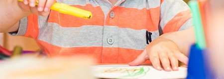Child holds a yellow marker and draws on paper, seated at a table; striped orange-gray polo shirt visible; blurred craft supplies and indoor classroom setting in the foreground.