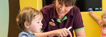 Toddler watches caregiver point at paper with a pen, learning at a table in a colorful playroom; another child sits in the background.
