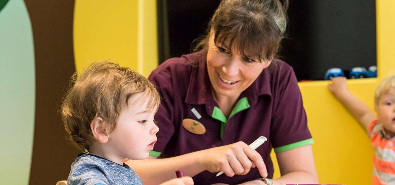 Adult caregiver guides a toddler drawing with crayons, pointing at paper. Context: bright childcare room with yellow walls, play alcoves, and another child watching in the background.