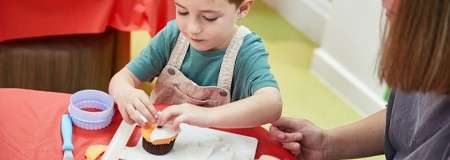 Child decorates a cupcake, placing orange icing on top, while an adult guides beside them; baking tools and cutters rest on a table covered with red cloth in a room.