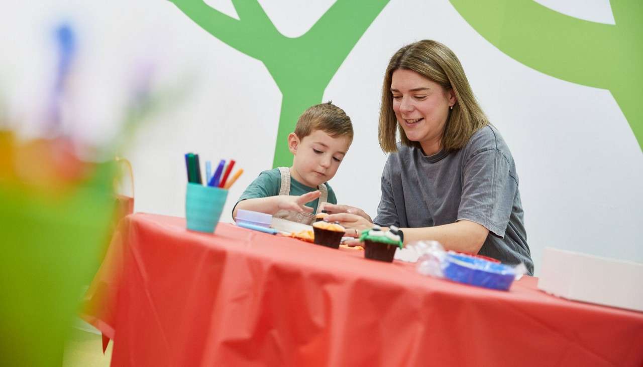 Child and adult decorate cupcakes, placing toppings by hand at a red-tablecloth craft table. Colored markers in a cup; supplies scattered. Background shows green tree shapes on a white wall.