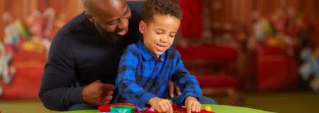 Child builds with colorful plastic pieces on a round green table, while an adult leans close, guiding. Background shows a warmly lit room with blurred red couches and toys.