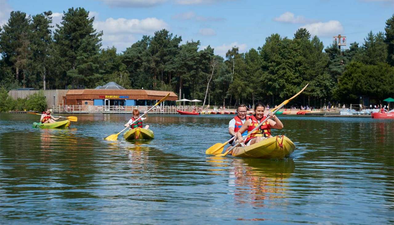 Kayakers paddle yellow canoes toward the camera, wearing life jackets. Behind them, a lakeside building reads "THE PANCAKE HOUSE," with docks, boats, and tall trees under a sunny sky.