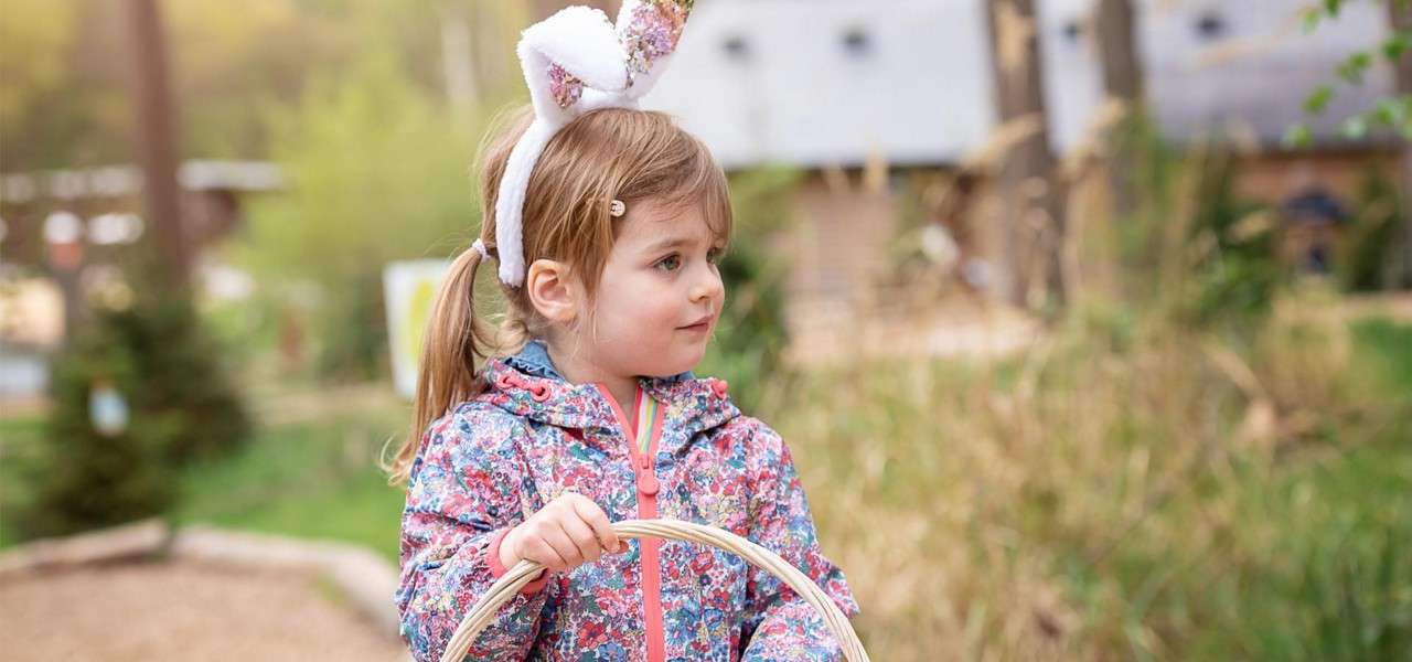 Young child wearing a bunny-ear headband holds a wicker basket, standing in a floral jacket; context: blurred outdoor park with trees, grass, and buildings in soft daylight.