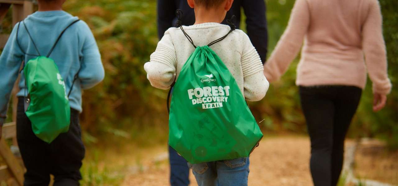 Child with a green drawstring backpack walks ahead along a wooded trail with companions. Backpack text reads: “Center Parcs FOREST DISCOVERY TRAIL.”