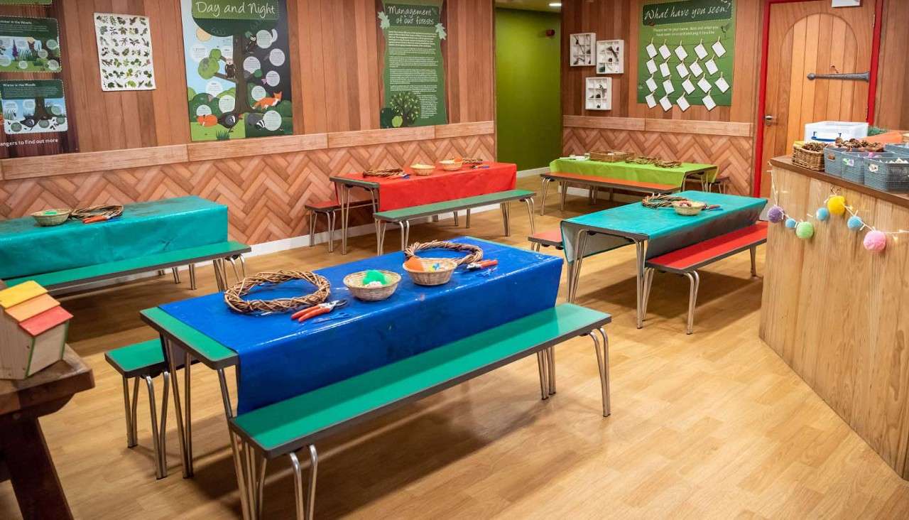 Craft tables display wreaths, baskets, and scissors, awaiting activities in a wood‑paneled classroom with benches and educational displays. Visible text: “Day and Night”; “Management of our”; “What have you seen?”