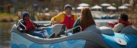 Four people converse in a blue pedal boat, wearing life jackets; the boat drifts on a calm park pond with reeds and patio umbrellas in the background. Text: “8080”, “1”.