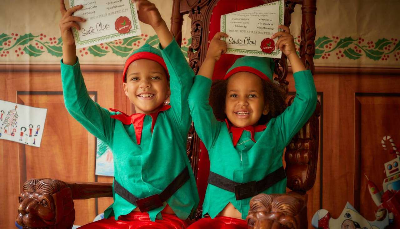 Two smiling children raise certificates while seated on carved wooden chairs, dressed as elves in a festive workshop backdrop with garlands and drawings. Certificate text (legible): “Santa Claus.” Remaining small print and red seal text are unreadable.