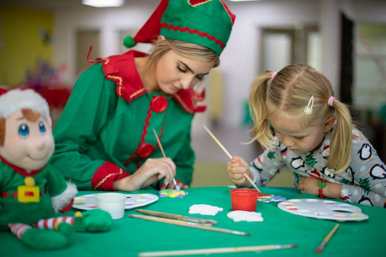 Two smiling children raise certificates while seated on carved wooden chairs, dressed as elves in a festive workshop backdrop with garlands and drawings. Certificate text (legible): “Santa Claus.” Remaining small print and red seal text are unreadable.