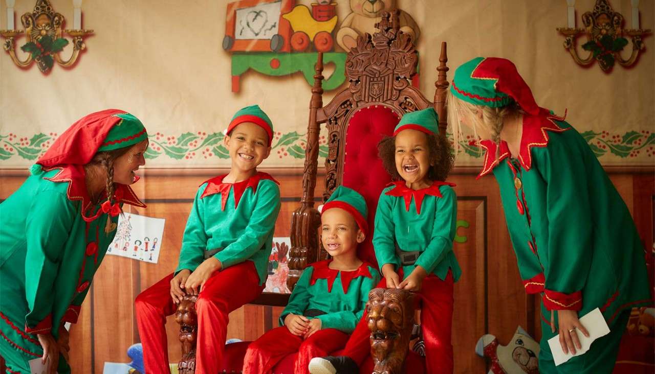 Children in elf costumes laugh and sit on a carved red throne while two adult elves lean in within a Christmas workshop set decorated with garlands, candles, and toy-train art.