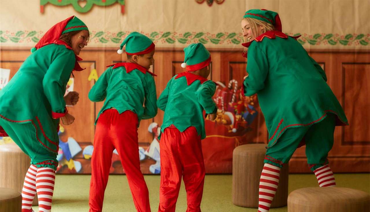 Four people dressed as Christmas elves bend and dance together, facing away. They wear green tunics, red pants, striped socks. Context: festive indoor set with wooden panels, candy-cane decorations, and small stools.