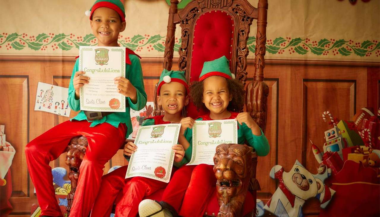 Three children dressed as elves hold certificates and smile, sitting on an ornate red velvet throne in a Christmas-decorated room with toys. Certificates text: “ACADEMY,” “Congratulations,” “Santa Claus.”