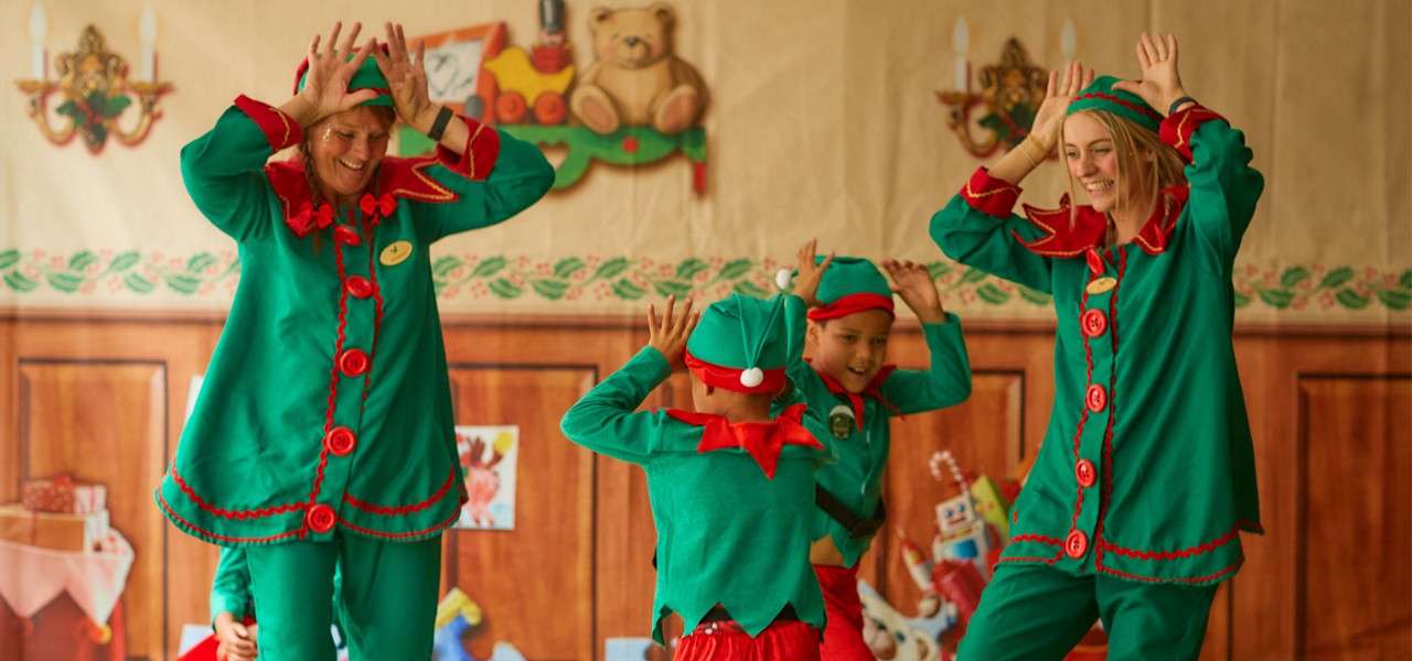 Four people dressed as Christmas elves dance with hands on heads in a festive room, wood-paneled backdrop with toys, wreaths, candles, and garlands.