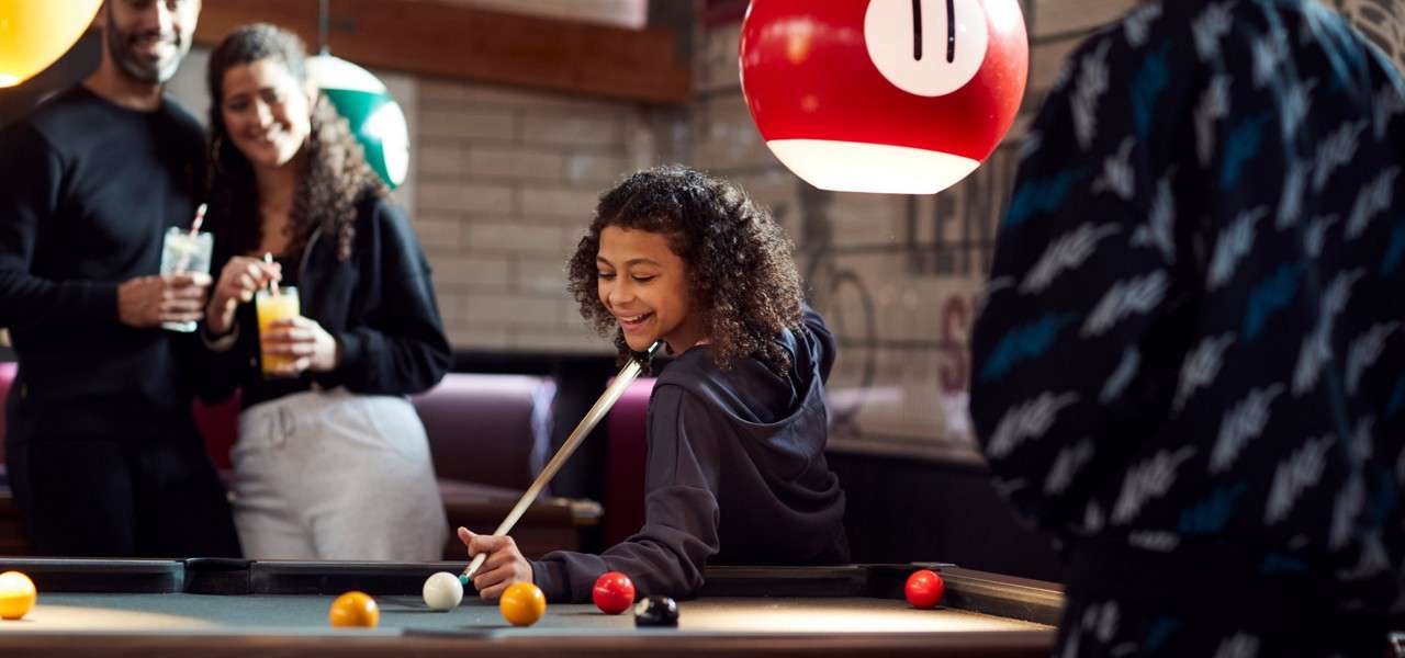 Girl lines up a shot on a pool table, smiling, while friends watch with drinks in a casual bar; an overhead red lamp marked 11 hangs above, balls scattered across the felt.