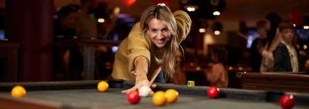 A person leans over a pool table, aiming a cue to strike the white ball; red and yellow balls scattered; dimly lit bar setting with people and warm lights in the background.