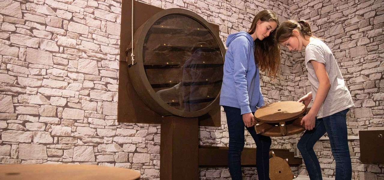 Two children examine and assemble round wooden pieces, collaborating beside a large circular wall mechanism; context: a stone-patterned room resembling an escape-room puzzle environment, with shelves and ropes nearby.