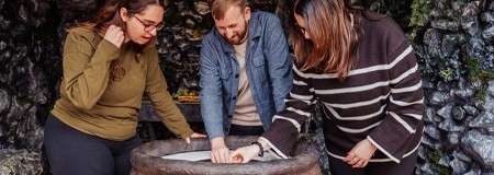 Three adults examine a paper on a round stone basin, leaning in and pointing; context: a dim, rocky cave-like room with mossy walls and a small table behind.
