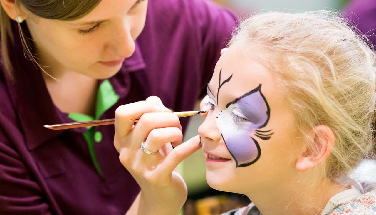 Child’s face—eyes closed—receives face paint; artist applies details with thin brush. Context: indoor event or festival, kid’s cheek decorated as purple butterfly with black outlines.