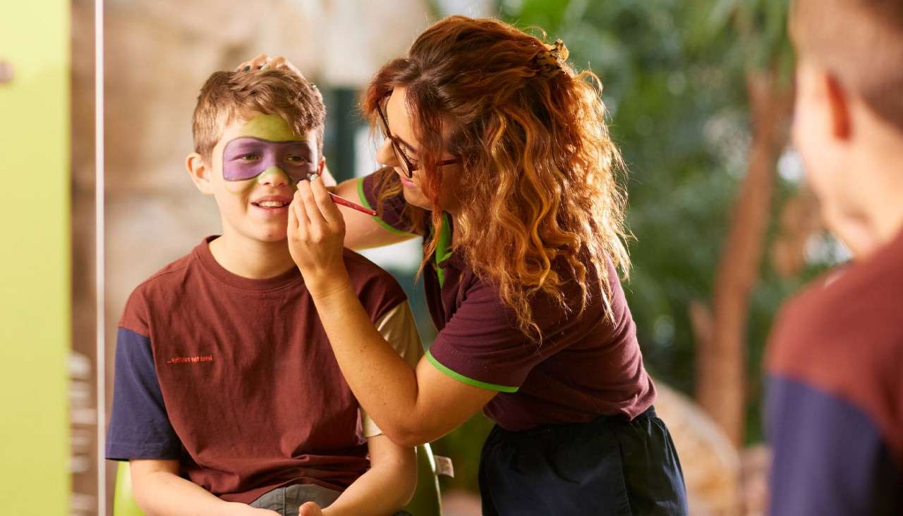 Face painter applies purple-and-green mask makeup to a smiling boy while another child watches; seated indoors with soft light, blurred greenery, and casual clothing suggesting a playful art activity.